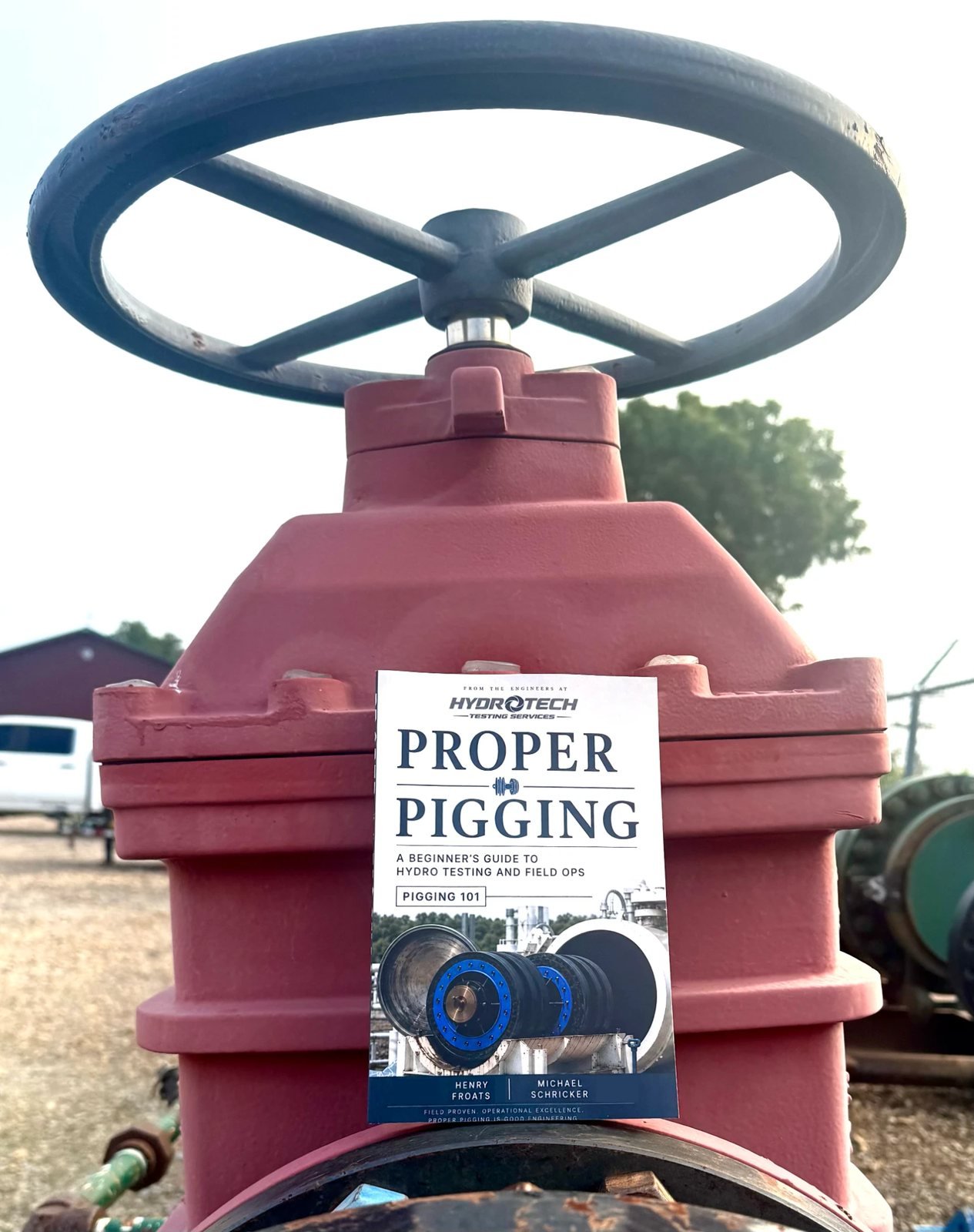 The printed Proper Pigging field guide by Henry Froats Jr. and Michael Schricker, propped against the bonnet of a red pipeline gate valve with its handwheel above, at a hydrostatic testing yard.