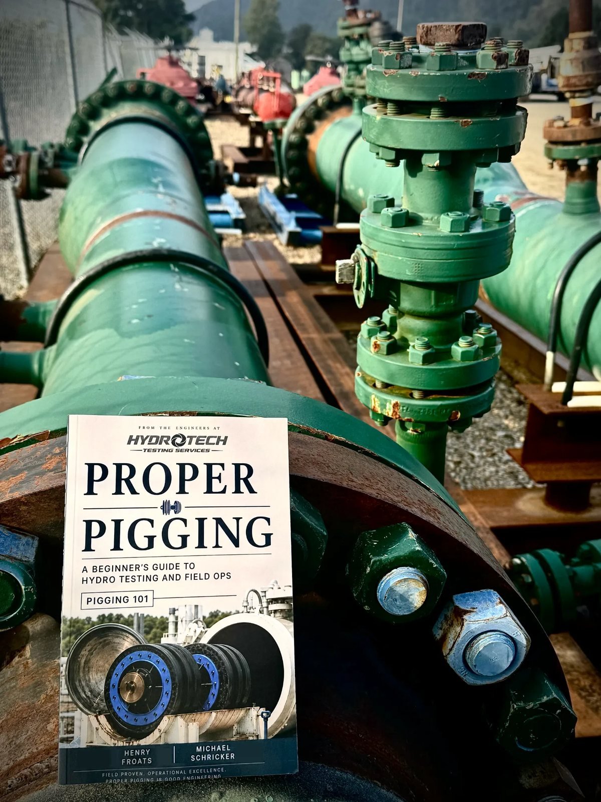 The printed Proper Pigging field guide by Henry Froats Jr. and Michael Schricker, standing on the flange face of a green-painted pipeline at a live hydrostatic test spread.